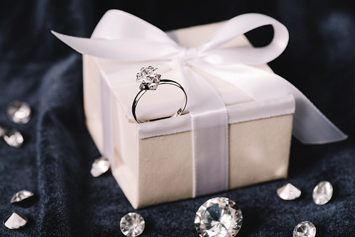 selective focus of engagement ring on gift box with bow near shiny diamonds on blue cloth