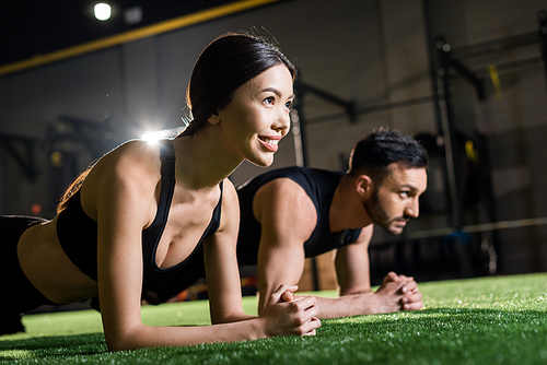 selective focus of cheerful woman smiling while doing plank exercise near man on grass