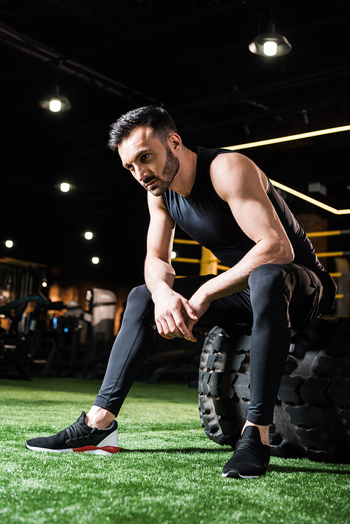 low angle view of handsome and tired athletic man sitting on car tire in gym