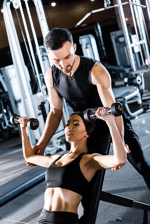 selective focus of young athletic woman working out with dumbbells near handsome  trainer in gym