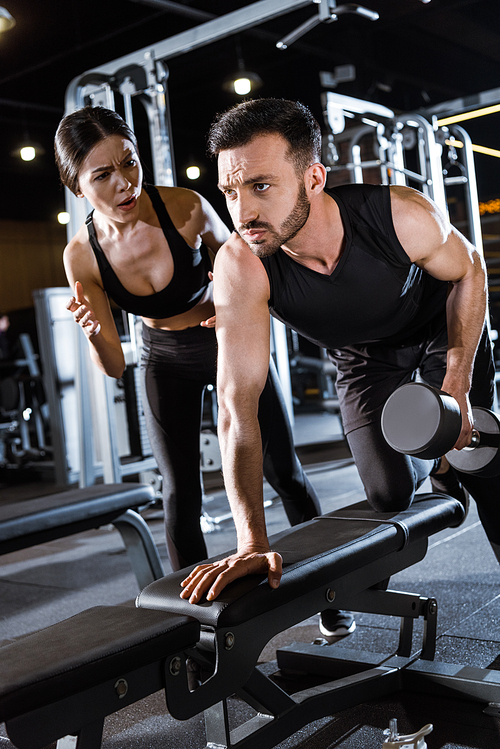 low angle view of attractive trainer standing near handsome man working out with dumbbell