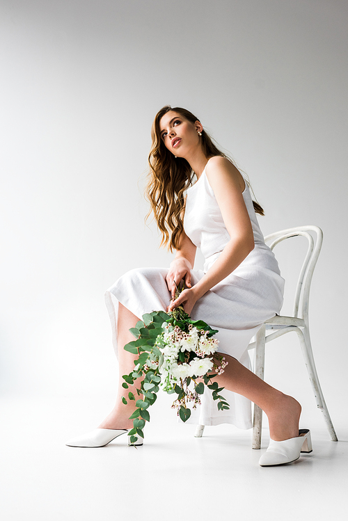 low angle view of woman in dress sitting on chair and holding bouquet of flowers with eucalyptus leaves on white