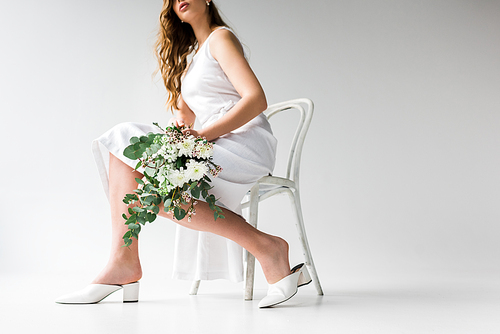 cropped view of woman in dress sitting on chair and holding bouquet of flowers with eucalyptus leaves on white
