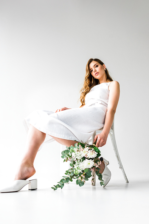 low angle view of young woman in dress sitting on chair and holding bouquet of flowers with eucalyptus leaves on white