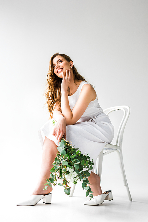happy young woman in dress sitting on chair and holding bouquet of flowers with eucalyptus leaves on white