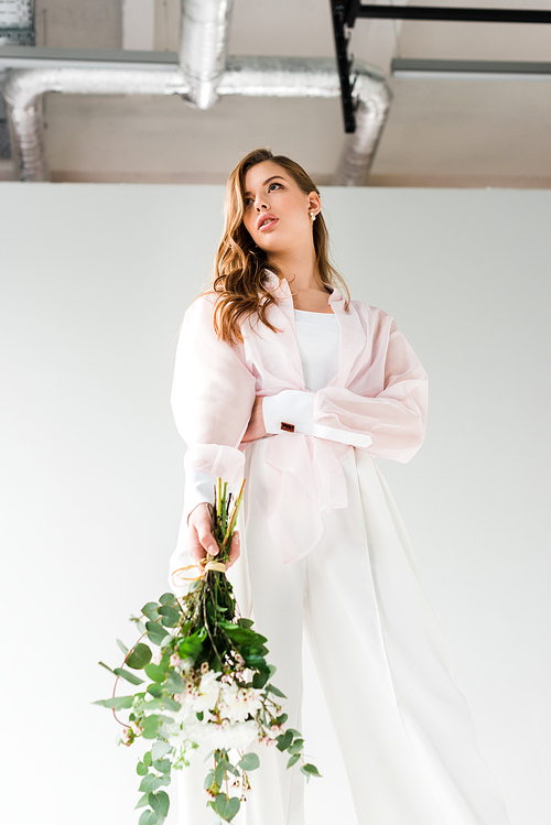 low angle view of attractive woman holding flowers with green eucalyptus leaves while standing on white