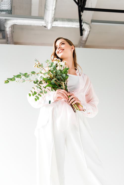 low angle view of happy woman holding flowers with green eucalyptus leaves while standing on white