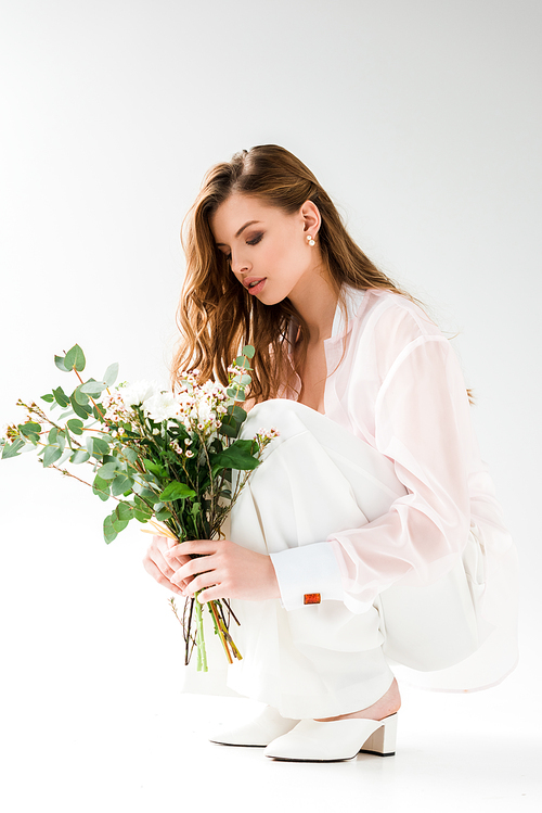 attractive woman looking at flowers with green eucalyptus leaves while sitting on white