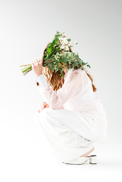 girl covering face with flowers with green eucalyptus leaves while sitting on white