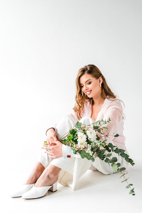smiling young woman sitting with flowers and green eucalyptus leaves on white