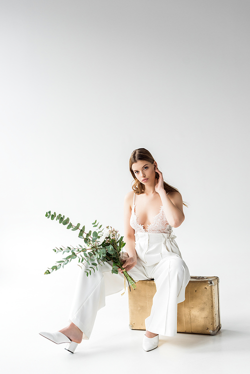 stylish young woman sitting on travel bag and holding bouquet with flowers and eucalyptus  leaves on white