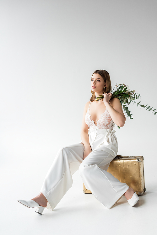 young woman sitting on travel bag and holding bouquet with flowers and eucalyptus leaves on white