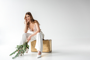 pensive girl sitting on travel bag and holding bouquet with flowers and eucalyptus leaves on white