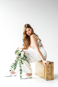 attractive girl sitting on travel bag and holding bouquet with flowers and eucalyptus leaves on white