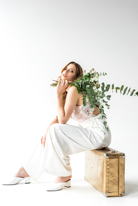 trendy girl sitting on travel bag and holding bouquet with flowers and eucalyptus leaves on white