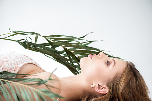 side view of attractive young woman lying near tropical palm leaves on white