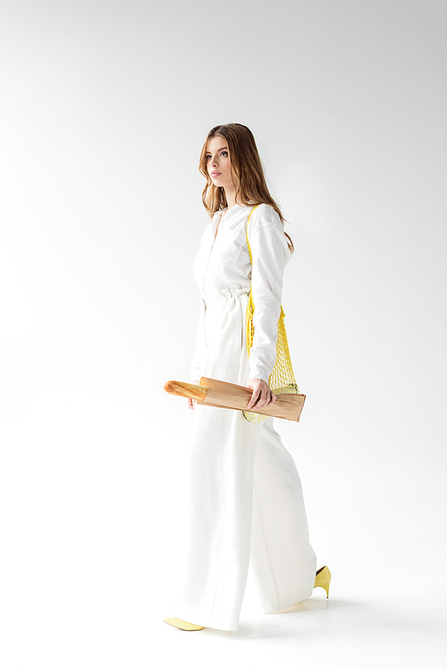 attractive woman walking with baguette in paper bag and yellow string bag on white
