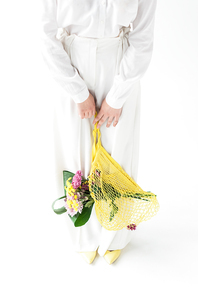 cropped view of woman holding yellow string bag with wildflowers while standing on white
