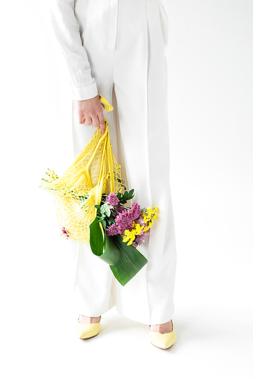 cropped view of girl holding yellow string bag with wildflowers while standing on white