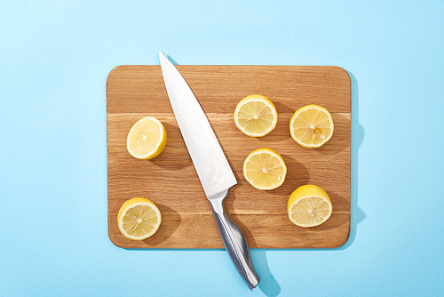 top view of ripe yellow cut lemons on wooden cutting board with knife on blue background
