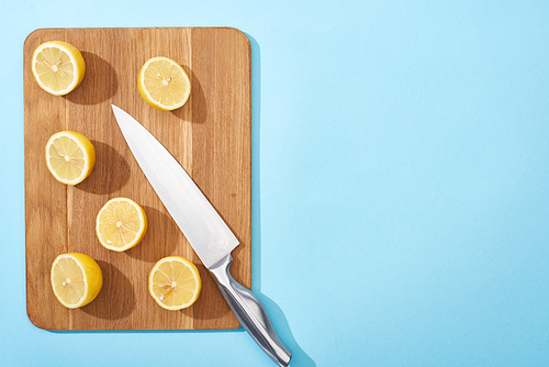 top view of ripe yellow cut lemons on wooden cutting board with knife on blue background with copy space