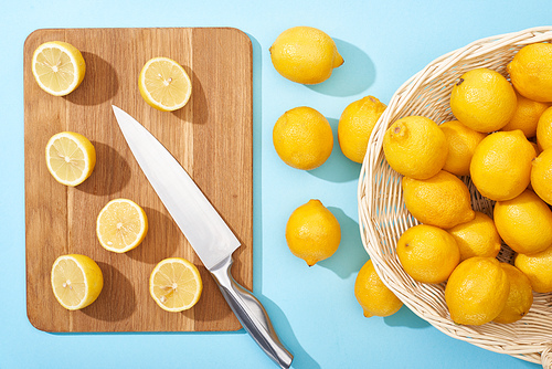 top view of ripe yellow cut lemons on wooden cutting board with knife on blue background near whole lemons in wicker basket