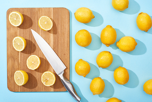 top view of ripe yellow cut lemons on wooden cutting board with knife on blue background near whole lemons
