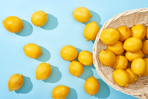 top view of ripe yellow lemons scattered from wicker basket on blue background