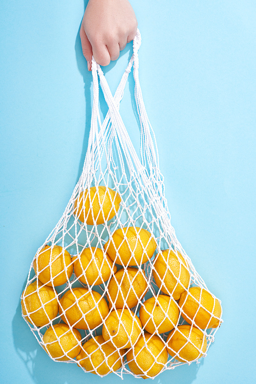 cropped view of woman holding ripe yellow lemons in string bag on blue background