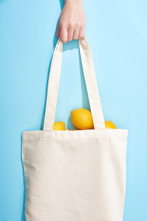 cropped view of woman holding ripe yellow lemons in cotton bag on blue background