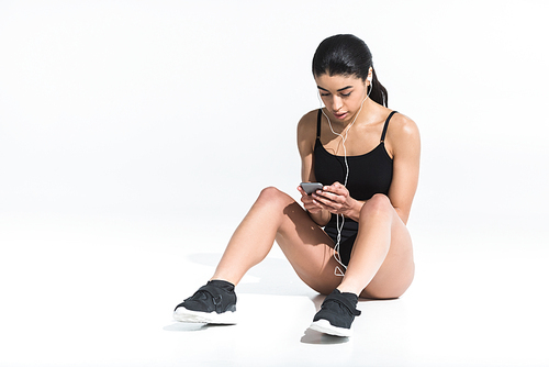 pretty african american girl in sportswear sitting on floor and listening music in earphones on white