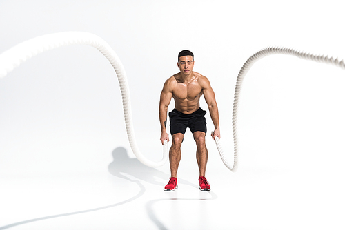 selective focus of handsome mixed race man in black shorts and red sneakers training with ropes on white