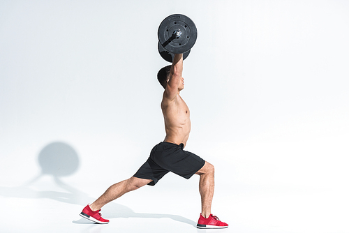 side view of athletic mixed race man in black shorts and red sneakers lifting barbell on white
