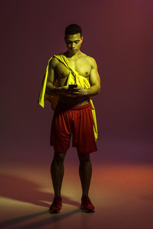 handsome mixed raxe man in red shorts and sneakers using smartphone on dark background