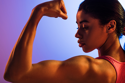 beautiful athletic african american girl demonstrating biceps on purple and blue gradient background