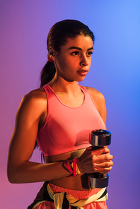 beautiful african american girl in pink sports bra holding dumbbell on purple and blue gradient background