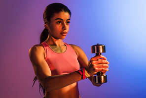 attractive african american woman in pink sports bra holding dumbbell on purple and blue gradient background