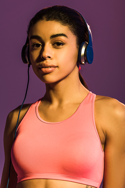 beautiful african american girl in pink sports bra and headphones  isolated on purple