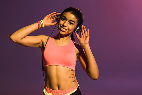sportive african american girl in pink sports bra and headphones smiling and  on purple background