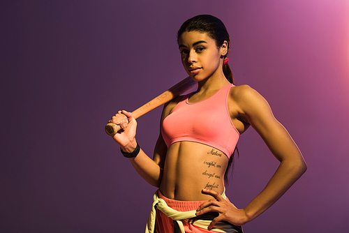 attractive african american girl with baseball bat posing at camera with hand on hip on purple background