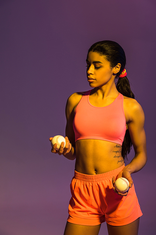 attractive sportive african american girl in pink sports bra and shorts holding balls on purple background