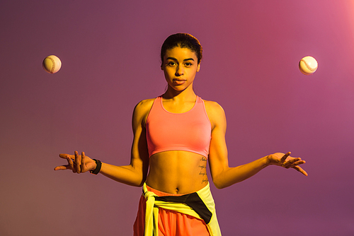 attractive african american girl in pink sports bra and shorts juggle with balls on purple background with gradient