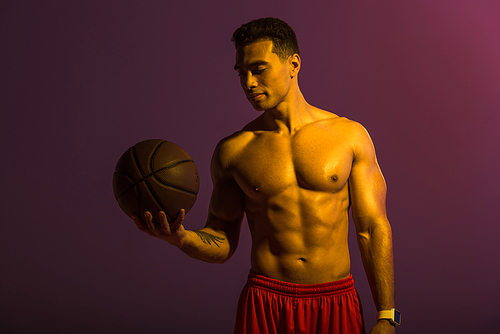 handsome athletic mixed race man holding brown ball on purple background