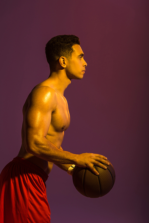 sportive shirtless mixed race man with muscular torso holding brown ball on purple background