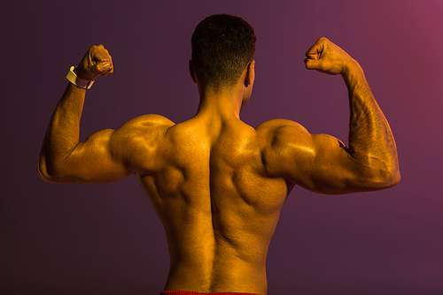 back view of athletic man with muscular torso demonstrating biceps on purple background