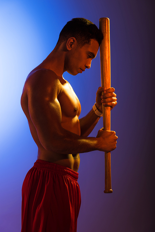 handsome mixed race man with muscular torso holding baseball bat on blue and dark purple gradient background