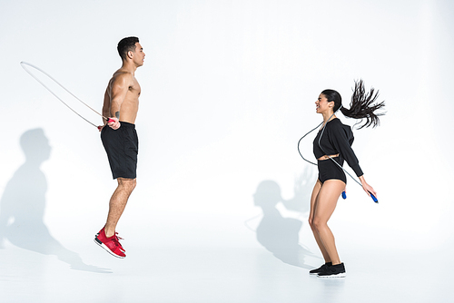 smiling african american woman and athletic mixed race man training with jump ropes on white