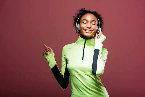 beautiful smiling african american sportswoman in headphones listening music with eyes closed isolated on brown