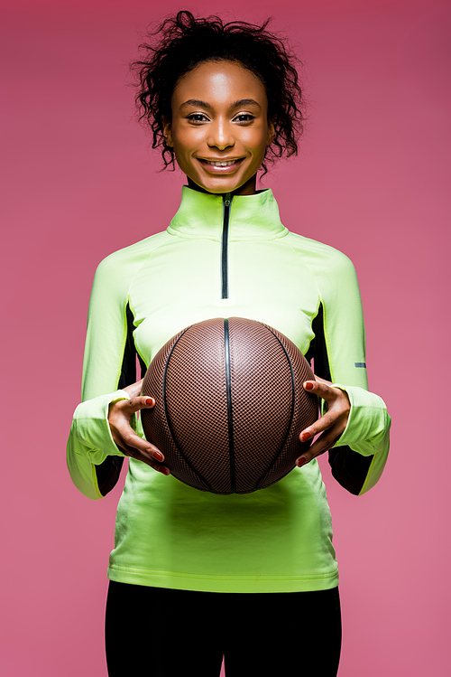 beautiful smiling african american sportswoman  and holding basketball isolated on pink