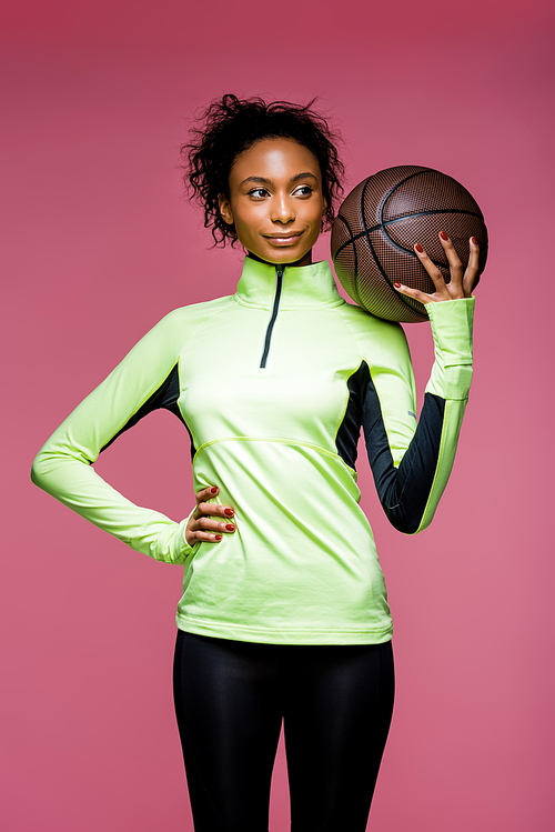 beautiful african american sportswoman with hand on hip holding basketball isolated on pink